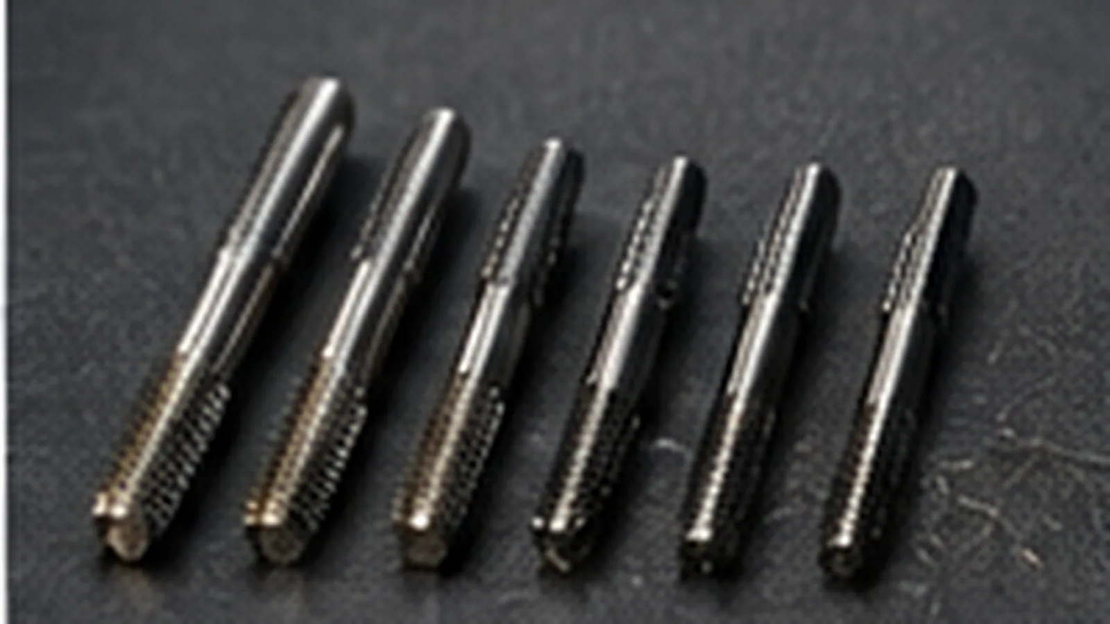 Six tarauds machine de diamètres croissants alignés sur un plan sombre, goujures et filets visibles.